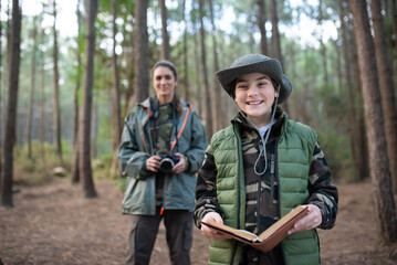 Cute little boy with notebook in forest. Schoolboy in coat and panama smiling at camera. Blurry mother in background. Childhood, nature, leisure concept