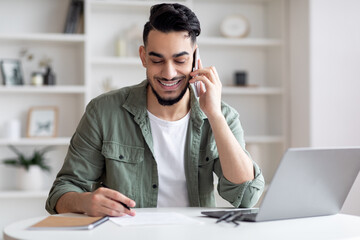 Handsome Arab Male Freelancer Talking On Cellphone And Taking Notes At Home