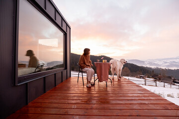 Woman with her dog resting on terrace of tiny house in the mountains, enjoying beautiful landscape...