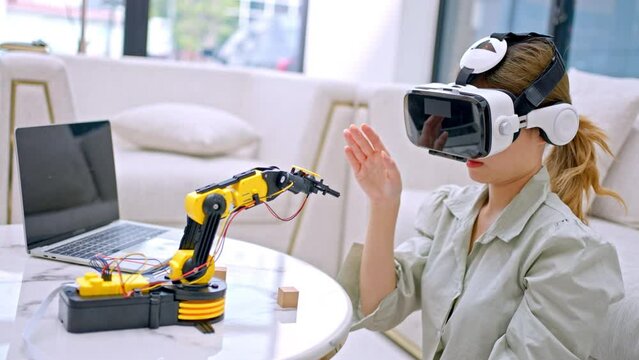 Engineer wearing (VR)virtual reality headset and gesturing while sitting at home.engineer is working on innovative technology mechanical Robot arm,She using laptop controlling Robotic arm by her hand.