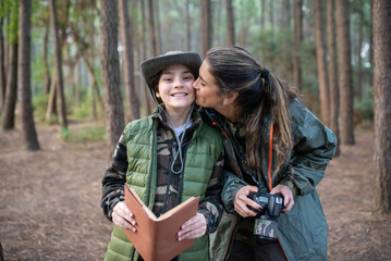 Loving mother with son with camera in forest. Female model in sportive clothes kissing smiling boy on cheek. Hobby, photography concept