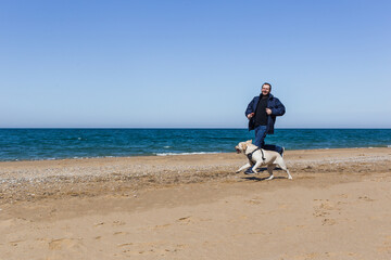 A man in winter clothes runs along a sandy beach with a dog in ammunition