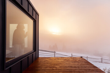 Tiny house with terrace in the mountains during winter on sunrise. Woman looks out from the window. Concept of small modern cabins for rest and escape to nature. Idea of solitude