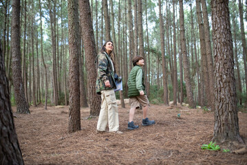 Content boy and woman with cameras walking in forest. Dark-haired mother and son in coats looking around. Parenting, family, leisure concept
