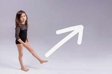 Strong little toddler girl raises her leg on a gray background. White arrow For women's empowerment, female power and feminism, sports, education and creative ideas of the future. Copy and space.