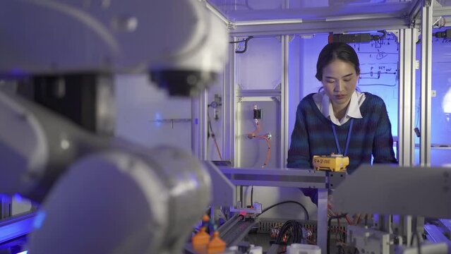 Panning left to right, selective focus of a young Asian female apprentice engineer using a digital multimeter checking an industrial machine voltage, with a blurred robotic arm machine in foreground.