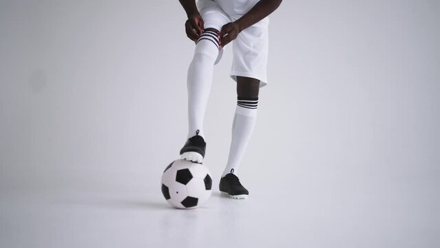 young sporty man is correcting his football uniform and holding ball by leg in studio