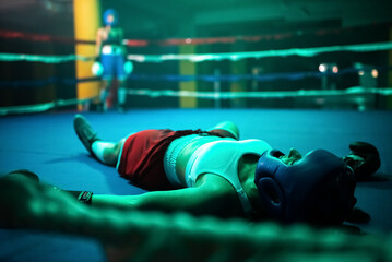 Close-up of boxer girl in knockout on ring. Two young girls boxing in gym when one girl fell down after took several blows of partner and lying on floor. Active lifestyle, hobby, womens boxing concept
