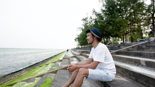 4K 50fps, Good looking Asian guy, sitting alone on a beautiful beach, enjoying the cool breeze, popular tourist destination