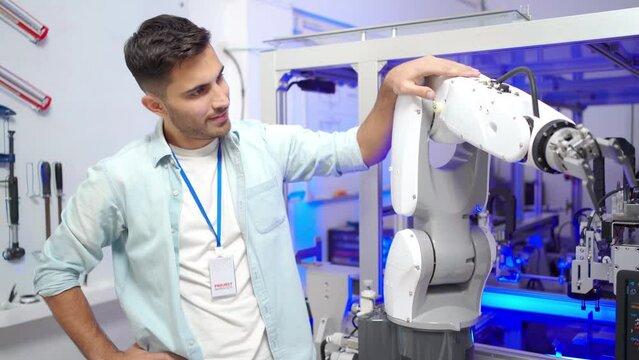 Young Caucasian male industrial engineer in blue shirt standing touching an automation robotic arm that are moving, then turn to looking at camera with arms folded and smiling in an industrial factory