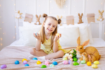 easter, a little girl with painted colored eggs, tulips and a rabbit at home in a bright room is preparing for the holiday smiling, having fun and playing with eggs closing her eyes