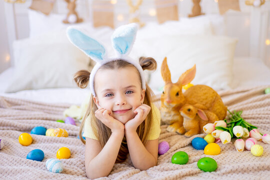 easter, a little child girl among painted colored eggs in bunny ears is preparing for the holiday smiling and having fun