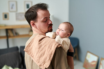 Young father looking at adorable baby son in romper suit on his shoulder covered with napkin while...