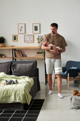 Young careful father in casualwear moving along comfortable double bed with toys while holding his adorable baby son on hands