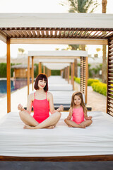 Smiling peaceful mom and daughter meditating at luxury sea resort sitting in lotus pose on gazebo near pool keeping zen hands smiling looking at the camera. Mom and 4s girl doing yoga, mental exercise