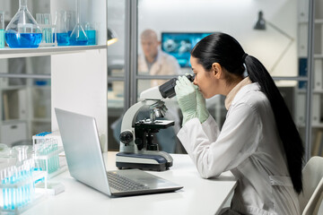 Side view of young female chemical researcher working with microscope while studying new substances by workplace in laboratory