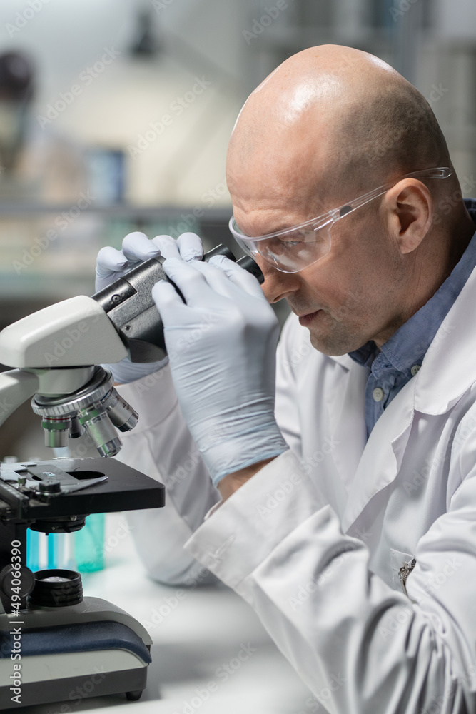Aufkleber Side view of bald mature man in whitecoat, gloves and eyeglasses using microscope while studing new chemical substance in laboratory