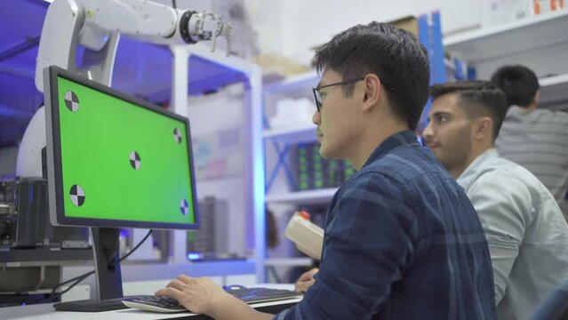 Selective focus, oblique angle of the back of Asian male data engineer sitting at desk keying a software code on a mock-up green screen, with a blurred Caucasian colleague using a controller near him.