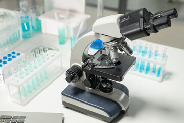 Microscope surrounded by groups of flasks containing blue liquid chemical substance standing on desk in laboratory
