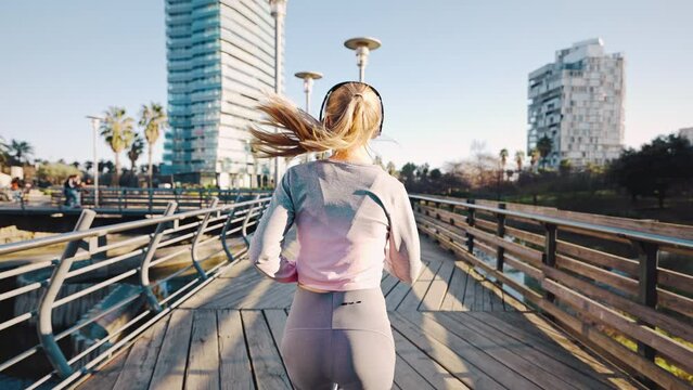 Camera follow from behind young Caucasian woman in tracksuit and sneakers running across big city bridge by music rhythm in earphones having everyday cardio workout training. Back view. Slow motion