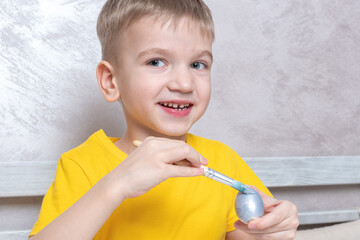 A little blond boy paints eggs for the Easter holiday at home. The child has fun and celebrates the holiday. DIY Easter eggs concept.