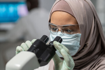Close-up of young Muslim female researcher looking in microscope during scientific investigation or...