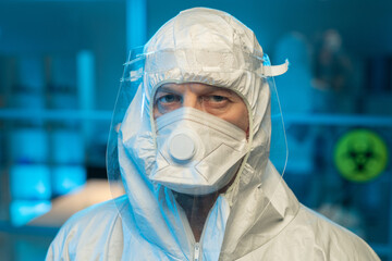 Fototapeta na wymiar Face of male researcher in biohazard suit, respirator and protective screen looking at camera against interior of laboratory