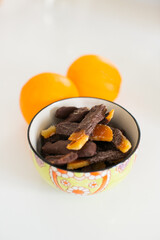 Pieces of chocolate dipped candied orange peel in small dish with oranges in the background