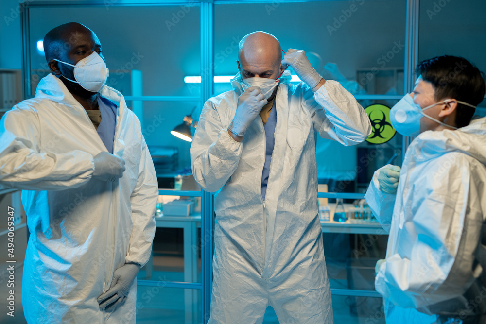 Poster Three interracial scientists putting on protective coveralls and respirators while going to work with biohazard substances in laboratory