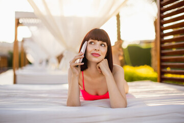 Summer vacations at luxury tropical resort. Happy smiling young woman, lying in wonderful wooden gazebo near the pool, talking to friend by phone. Call from paradise. Gazebo row on background.