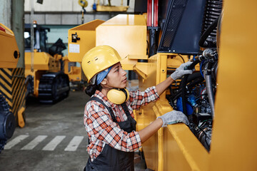 Young Hispanic female in protective helmet, coveralls and gloves checking engine of huge industrial...