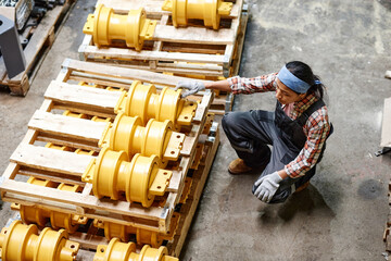 Above angle of young female worker of industrial plant sitting by stack of spare parts while...