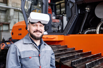 Happy mature worker of factory wearing vr headset and workwear looking at cameras while standing by...