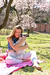 Panoramic view of woman hugging dog on picnic blanket at park in springtime. Vertical full view of young woman in love with french bulldog at park in summer. Animals and people lifestyle.