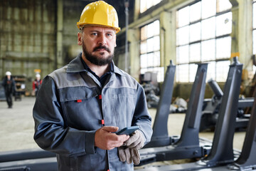 Serious mature workman or technician with smartphone in hardhat and boilersuit standing in large distribution warehouse