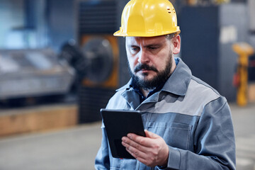 Mature bearded male worker of industrial plant in boilersuit and hardhat looking through data on screen of digital tablet