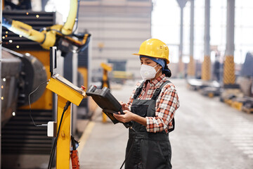 Young female technician in protective workwear using remote switch to control industrial machine while standing in large workshop