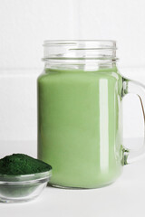 fresh green smoothie in glass bottle and spirulina powder with spoon on white background. useful habits, self care and healthy lifestyle. copy space