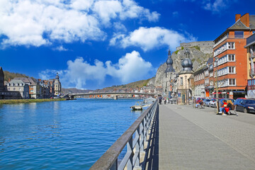 Dinant, Belgium - March 9. 2022: View on meuse river bank urban promenade with bridge, church, rock face against blue sky with white fluffy clouds