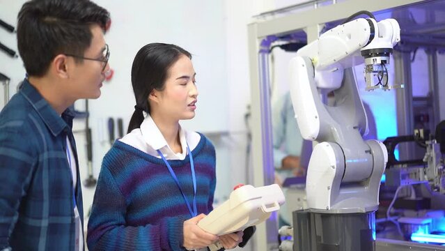 Asian female apprentice engineer in student uniform testing the work of an automation robotic arm machine with a robot controller with a young Asian male CEO giving an advice next to her in factory.