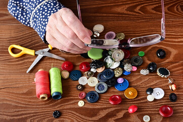elderly woman looks through glasses at colorful buttons on wooden table. Sewing accessories for needlework, hobbies and handmade