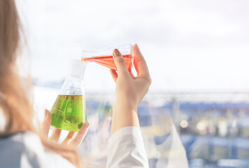 A girl laboratory assistant standing at the window mixes reagents in flasks in the laboratory at...