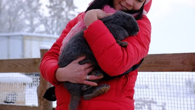 An adult woman hugs a big black rabbit. Farming, veterinary. Pet. A woman in a red jacket hugs an animal in the street with love.
