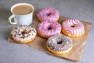 Several donuts lie on craft parchment paper on gray surface. Porcelain cup of coffee stands next to donuts laid out on table. Selective focus.
