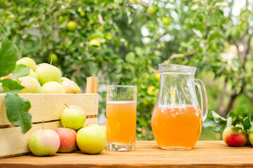 A box of apples and glass and pitcher of fresh apple juice on wooden table with garden background....