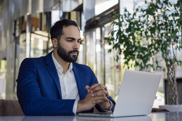 Handsome moroccan business man using laptop computer watching training courses working online in modern office. Young pensive middle eastern manager planning project sitting at workplace 