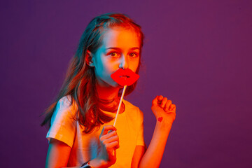 Portrait of a child girl. The girl put paper lips to her lips on a purple background.