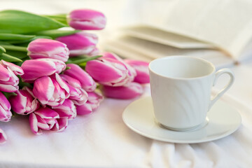 Fototapeta na wymiar A bouquet of beautiful pink tulips lies on a white bed, next to a white cup and saucer and an open book