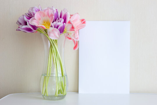 A bouquet of open pink tulips in a glass vase stands on a white table against a light wall.