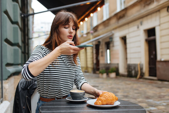 Woman taking photo of coffee and croissant on cell phone